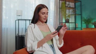 Portrait of lovely girl use mobile smartphone typing browsing say Wow yes found out great big win good news celebrate doing winner gesture. Young woman at home in living room sitting on orange sofa