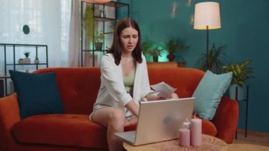 Stressed girl at home room looking at unpaid bank debt bills, doing paperwork, planning budget, calculate finances mortgage payments. Displeased young woman throws paper bills sitting on orange couch