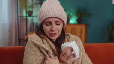 Sick girl wear hat wrapped in plaid sit alone shivering from cold on sofa drinking hot tea in unheated apartment without heating due debt. Unhealthy young woman feeling discomfort try to warming up