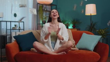 Money cash hundred euro rain falling on girl celebrating success, winning, goal achievement, good news, lottery jackpot luck at modern home office apartment indoors. Career grow. Woman sits on sofa