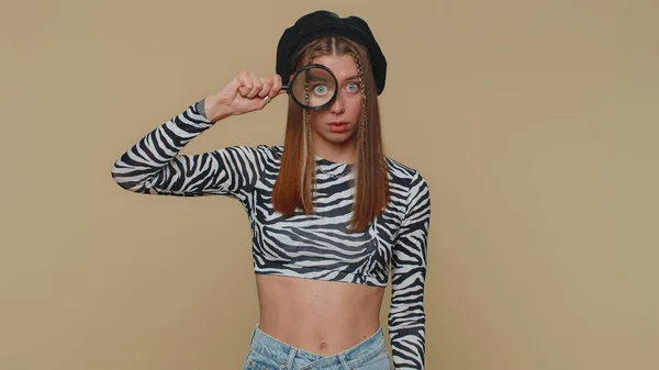 Investigator researcher scientist woman holding magnifying glass near face, looking into camera with big zoomed funny eye, searching, analysing. Adult stylish girl on beige studio background indoors