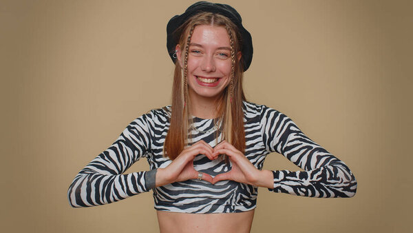 I love you. Smiling lovely pretty young woman makes heart gesture demonstrates love sign expresses good feelings and sympathy. Adult stylish female girl isolated on beige studio background indoors