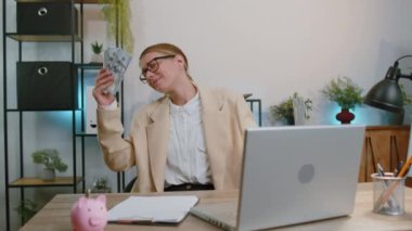Happy businesswoman working on laptop computer sincerely rejoicing win, receiving money dollar cash banknotes, success lottery luck at office workplace. Professional manager freelancer woman dancing