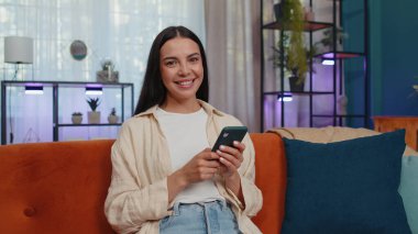 Portrait of adult girl sitting on sofa uses mobile phone smile at modern home apartment. Young woman texting share messages content on smartphone social media applications online, watching relax movie