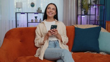 Portrait of adult girl sitting on sofa uses mobile phone smile at modern home apartment. Young woman texting share messages content on smartphone social media applications online, watching relax movie