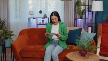 Stressed girl at home room looking at unpaid bank debt bills, doing paperwork, planning budget, calculate finances mortgage payments. Displeased young woman with paper bills sitting on orange couch