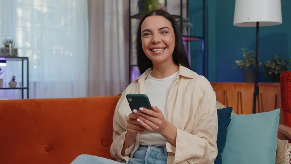 Portrait of adult girl sitting on sofa uses mobile phone smile at modern home apartment. Young woman texting share messages content on smartphone social media applications online, watching relax movie