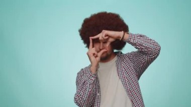Photographer man gesturing picture frame with hands, looks through fingers and focusing on interesting moment, imitating zoom and cropping nice image. Young guy isolated on blue studio background