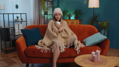 Sick girl wear hat wrapped in plaid sit alone shivering from cold on sofa drinking hot tea in unheated apartment without heating due debt. Unhealthy young woman feeling discomfort try to warming up
