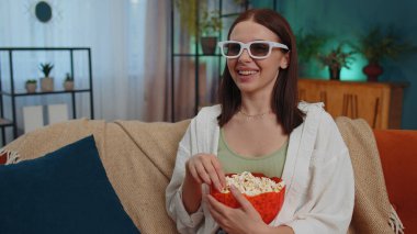 Excited adult girl sitting on sofa eating popcorn and watching interesting tv serial, sport game, film, online social media movie content online at home. Young woman enjoying domestic entertainment