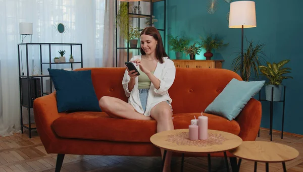 Portrait of adult girl sitting on sofa uses mobile phone smile at modern home apartment. Young woman texting share messages content on smartphone social media applications online, watching relax movie