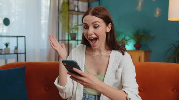 Oh my God Wow. Excited happy joyful winner girl use smartphone typing browsing found out great win good news celebrate victory. Young woman with mobile phone sitting at home in room on orange couch