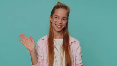 Wow. Hello, welcome. Lovely pretty teenager girl appear from below waving hi with her palm, greeting with hospitable friendly smile. Young female child kid isolated on blue studio background indoors