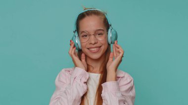 Happy pretty teenager relaxed girl in glasses listening music via headphones and dancing disco fooling around having fun expressive gesticulating hands. Young stylish teen female child kid indoors