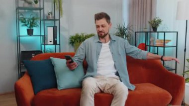 Portrait of adult man sitting on sofa uses mobile phone smiles at modern home apartment. Young guy texting share messages content on smartphone social media applications online, watching relax movie
