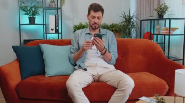 Planning family budget. Smiling happy man counting money dollar cash, use smartphone calculate domestic bills at home. Joyful young guy satisfied of income saves money for planned vacation, gifts