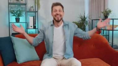 Happy caucasian man shouting, celebrating success, winning, goal achievement, good news, raising fists in gesture I did it, lottery luck at home apartment. Young guy indoors in living room on couch