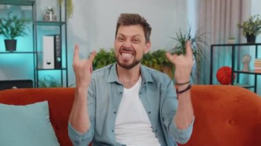 Overjoyed delighted caucasian adult man showing rock n roll gesture by hands, cool sign, shouting yeah with crazy expression, dancing, emotionally rejoicing in success. Young guy at home apartment