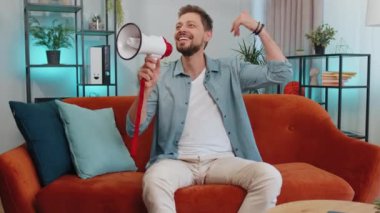 Portrait of adult man talking with megaphone, proclaiming news, loudly announcing advertisement, warning using loudspeaker to shout speech. Young guy at modern home apartment living room on couch