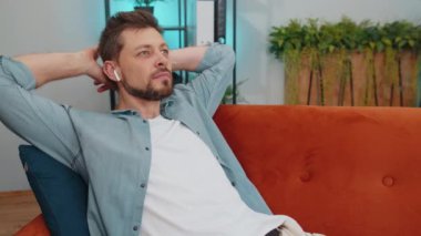 Young adult man in wireless earphones relaxing lying on sofa at home choosing listening favorite energetic disco rock n roll dancing music in smartphone. People weekend activities. Happy overjoyed guy