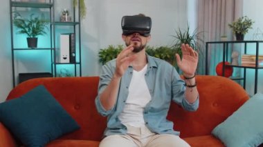 Young man using virtual reality futuristic technology VR app headset helmet to play simulation 3D 360 video game, watching film movie at modern home apartment. Adult guy in goggles sitting on couch