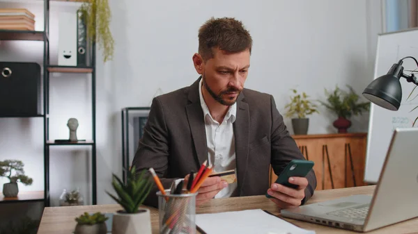 Businessman programmer software developer in suit working, making online purchase payment shopping with credit bank card and smartphone at modern office desk with laptop. Male freelancer business man