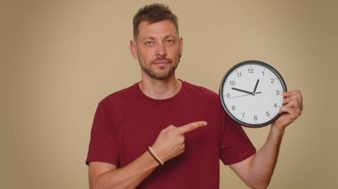 It is your time. Handsome young man in red t-shirt showing time on wall office clock, ok, thumb up, approve, pointing finger at camera. Adult stylish male guy on beige studio wall background indoor