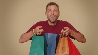 Happy young man showing shopping bags, advertising discounts, smiling looking amazed with low prices, shopping on Black Friday holidays. Adult stylish guy isolated alone on beige studio background
