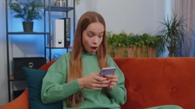 Teen girl use mobile smartphone typing browsing say Wow yes found out great big win good news celebrate lottery jackpot doing winner gesture. Young child kid at home in living room sitting on sofa