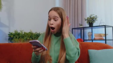Oh my God Wow. Excited happy joyful winner girl use smartphone typing browsing found out great win good news celebrate victory. Young child kid with mobile phone sits at home in room on orange couch