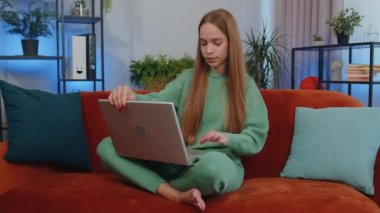 Lovely teen girl freelancer at home living room and sitting on orange couch sofa, opens laptop. Young child kid working on notebook, sends messages, makes online purchases, watching movies, working
