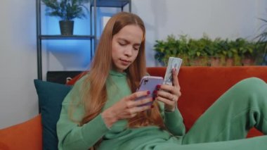 Planning family budget. Smiling happy girl counting money dollar cash, use smartphone calculate domestic bills at home. Young child kid satisfied of income saves money for planned vacation, gifts