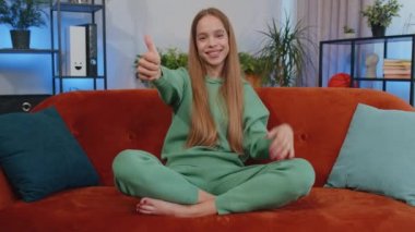 Like. Happy lovely teen girl looking approvingly at camera showing thumbs up, like sign positive something, good news, positive feedback. Young child kid sitting on sofa couch at home living room