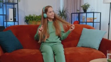Happy relaxed overjoyed child kid in wireless headphones dancing on couch at home choosing listening favorite energetic disco rock n roll music in smartphone entertaining. People weekend activities