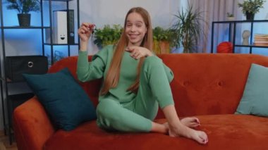 Young child kid real estate agent lifting hand showing the keys of new home house apartment, buying or renting property, mortgage loan at home apartment indoors. Lovely girl in room sitting on sofa