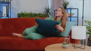 Repair work at neighbours concept. Irritated girl sit on couch cover ears with pillows annoyed by noisy neighbors suffer from headache wishes silence. Thin walls at home flat without sound insulation