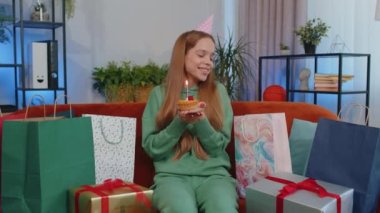 Happy teen girl wears festive birthday hat hold cupcake makes wish joyful congratulating blowing burning candle on cake. Young child kid celebrating anniversary party at home room apartment on couch