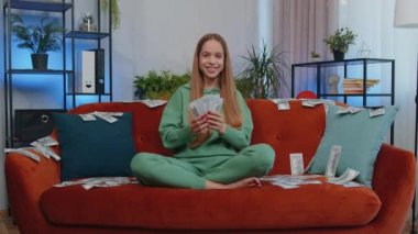 Money cash dollar rain falling on girl celebrating success, winning, goal achievement, good news, lottery jackpot luck at modern home office apartment indoors. Career grow. Young woman sits on sofa