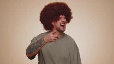 Amused happy man in casual t-shirt pointing finger to camera, laughing out loud, taunting making fun of ridiculous appearance, funny joke. Young guy boy posing alone on beige studio wall background