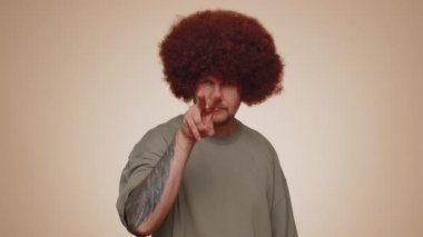 I am watching you. Man with lush hairstyle wig pointing at his eyes and camera, show I am watching you gesture, spying on someone. Young adult guy boy isolated alone on beige studio wall background