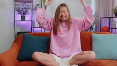 Happy caucasian girl shouting, celebrating success, winning, goal achievement, good news, raising fists in gesture I did it, lottery luck at home apartment. Young woman indoors in living room on couch