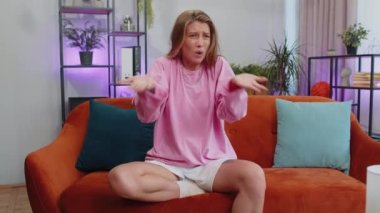 What. Why. Lovely girl raising hands in indignant expression, asking reason of failure, demonstrating disbelief irritation by troubles at home apartment. Young woman in room sitting on orange sofa