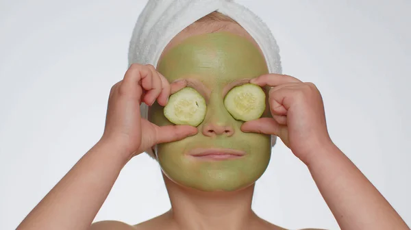 Smiling child girl applying cleansing moisturizing green facial mask, holding slices of cucumber, covering eyes. Teenager kid face skin care treatment natural cosmetics. Female close-up macro portrait