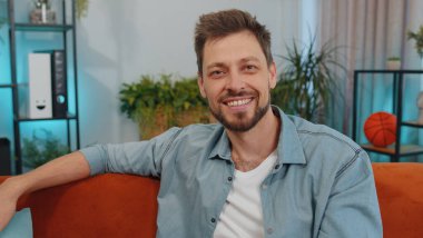 Close-up of happy calm adult man in shirt smiling friendly, glad expression looking away dreaming resting, relaxation feel satisfied concept good news. Young caucasian guy sitting on sofa at home room