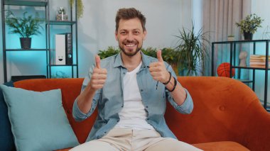 Like. Happy Handsome adult man looking approvingly at camera showing thumbs up, like sign positive something, good news, positive feedback. Young guy sitting on orange couch at home living room