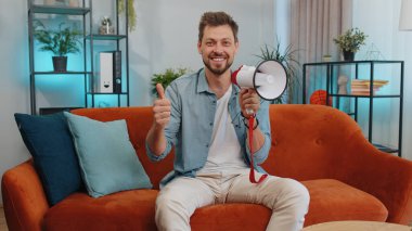 Portrait of adult man talking with megaphone, proclaiming news, loudly announcing advertisement, warning using loudspeaker to shout speech. Young guy at modern home apartment living room on couch