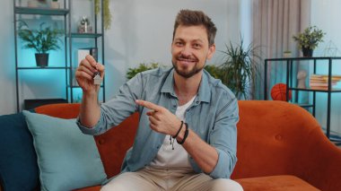 Young man real estate agent lifting hand showing the keys of new home house apartment, buying or renting property, mortgage loan at modern home apartment indoors. Handsome guy in room sitting on sofa