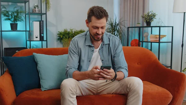 Portrait of adult man sitting on sofa uses mobile phone smiles at modern home apartment. Young guy texting share messages content on smartphone social media applications online, watching relax movie