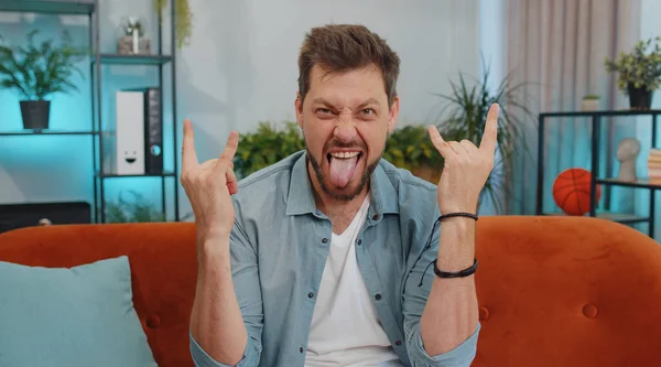 Overjoyed delighted caucasian adult man showing rock n roll gesture by hands, cool sign, shouting yeah with crazy expression, dancing, emotionally rejoicing in success. Young guy at home apartment