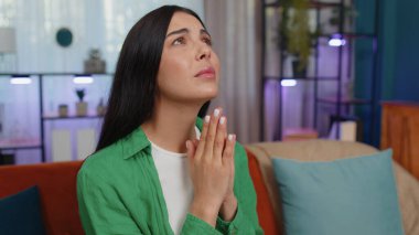 Please, God help. Girl praying sincerely with folded arms, looking upward and making wish, asking with hopeful imploring expression, begging apology. Young woman sitting on couch at home living room